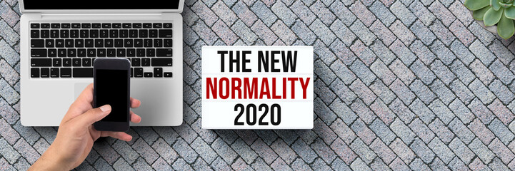 lightbox with message NEW NORMALITY 2020 with laptop and hand holding smartphone on stone pavement background