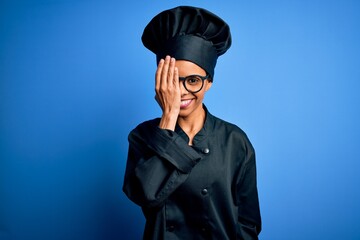 Wall Mural - Young african american chef woman wearing cooker uniform and hat over blue background covering one eye with hand, confident smile on face and surprise emotion.