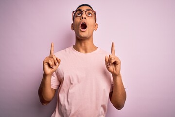 Wall Mural - Handsome african american man wearing casual t-shirt and glasses over pink background amazed and surprised looking up and pointing with fingers and raised arms.