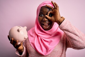 Poster - Young african american plus size woman wearing muslim hijab holding pink piggy bank with happy face smiling doing ok sign with hand on eye looking through fingers