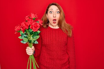 Poster - Young beautiful redhead woman holding bouquet of red roses flowers over isolated background scared in shock with a surprise face, afraid and excited with fear expression