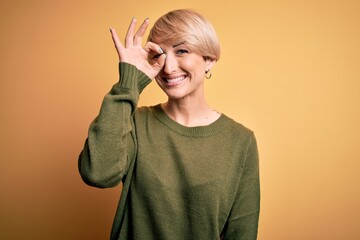 Canvas Print - Young blonde woman with modern short hair wearing casual sweater over yellow background doing ok gesture with hand smiling, eye looking through fingers with happy face.