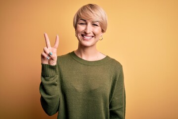 Poster - Young blonde woman with modern short hair wearing casual sweater over yellow background showing and pointing up with fingers number two while smiling confident and happy.