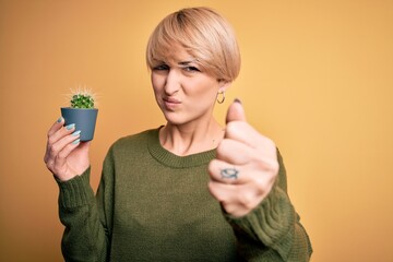 Sticker - Young blonde woman with short hair holding succulent green cactus over yellow background annoyed and frustrated shouting with anger, crazy and yelling with raised hand, anger concept