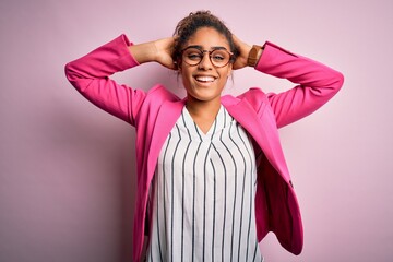 Wall Mural - Beautiful african american businesswoman wearing jacket and glasses over pink background relaxing and stretching, arms and hands behind head and neck smiling happy