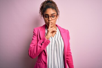 Poster - Beautiful african american businesswoman wearing jacket and glasses over pink background asking to be quiet with finger on lips. Silence and secret concept.