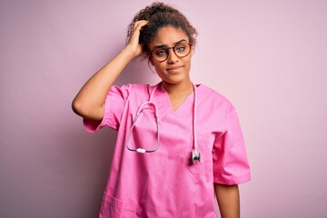 Wall Mural - African american nurse girl wearing medical uniform and stethoscope over pink background confuse and wonder about question. Uncertain with doubt, thinking with hand on head. Pensive concept.