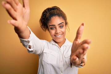 Poster - Young beautiful african american girl wearing casual shirt standing over yellow background looking at the camera smiling with open arms for hug. Cheerful expression embracing happiness.