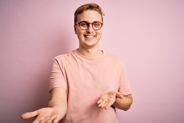 Wall Mural - Young handsome redhead man wearing casual t-shirt standing over isolated pink background smiling cheerful with open arms as friendly welcome, positive and confident greetings