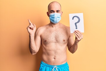 Sticker - Young handsome bald man wearing swimwear and medical mask holding question mark smiling happy pointing with hand and finger to the side