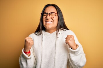 Poster - Young beautiful asian woman wearing casual sweater and glasses over yellow background very happy and excited doing winner gesture with arms raised, smiling and screaming for success. Celebration