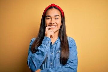 Poster - Young beautiful asian woman wearing casual denim shirt and diadem over yellow background looking confident at the camera with smile with crossed arms and hand raised on chin. Thinking positive.