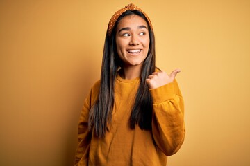 Poster - Young beautiful asian woman wearing casual sweater and diadem over yellow background smiling with happy face looking and pointing to the side with thumb up.