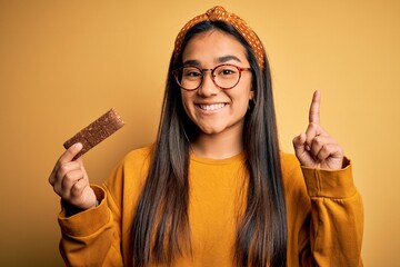 Poster - Young beautiful asian woman eating healthy protein bar over isolated yellow background surprised with an idea or question pointing finger with happy face, number one