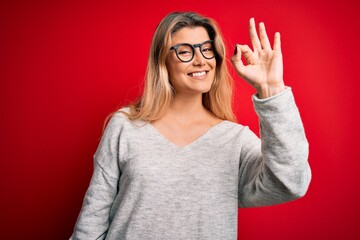 Poster - Young beautiful blonde woman wearing sweater and glasses over isolated red background smiling positive doing ok sign with hand and fingers. Successful expression.