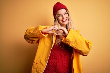 Sticker - Young beautiful blonde woman wearing raincoat and wool cap over isolated yellow background smiling in love showing heart symbol and shape with hands. Romantic concept.