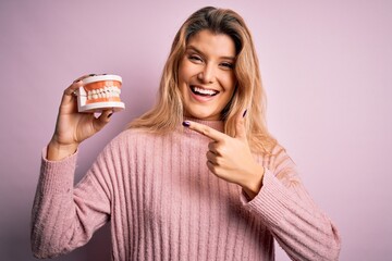 Sticker - Young beautiful blonde woman holding plastic denture over isolated pink background very happy pointing with hand and finger