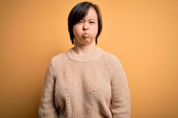 Wall Mural - Young down syndrome woman wearing casual sweater over yellow background puffing cheeks with funny face. Mouth inflated with air, crazy expression.