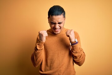 Wall Mural - Young brazilian man wearing casual sweater standing over isolated yellow background excited for success with arms raised and eyes closed celebrating victory smiling. Winner concept.