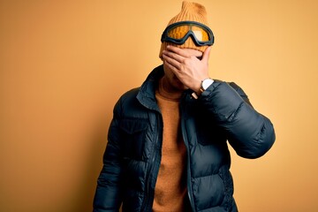 Wall Mural - Young brazilian skier man wearing snow sportswear and ski goggles over yellow background covering eyes with hand, looking serious and sad. Sightless, hiding and rejection concept