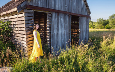 A close portrait of an Asian woman with long hair and yellow dress in front of an old barn on a farmland