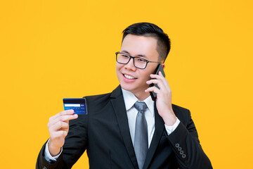 Portrait of smiling young Asian businessman having conversation on phone while holding and looking at credit card in isolated studio yellow background