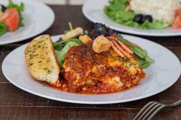 Piece of the delicious lasagna with garlic bread and veggies on a white plate with a  fork