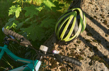 A light green bicycle helmet lies on a large stone in the park. Next to the grass lies a blue teenage bike. The concept of an active lifestyle and a safe ride.