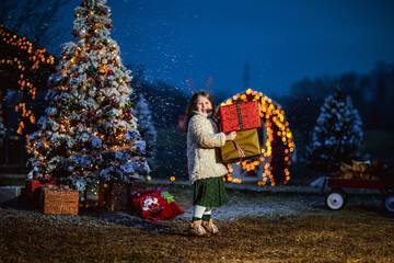 Cute girl with long curly hair in beige coat holding big presents against Christmas decorations. Copy space.