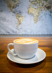 Coffee in white cup sitting on a wooden table with the world map hanging on the background. Wanderlust and hygge concept.