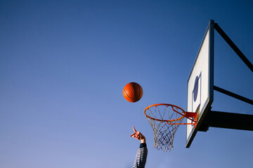 Wall Mural - Street basketball ball player throwing ball into the hoop. Close up of hand, orange ball above the hoop net with blue sky in the background. Concept of success, scoring points and winning. Copy space