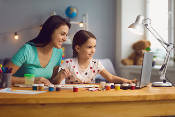 Happy mother helping her cute daughter study online at home. Parent and child watching art lesson together on laptop