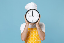 Young Bearded Male Chef Or Cook Baker Man In Apron White T-shirt Toque Chefs Hat Isolated On Pastel Blue Background Studio Portrait. Cooking Food Concept. Mock Up Copy Space. Covering Face With Clock.