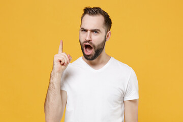 Displeased shocked young bearded man guy in white casual t-shirt posing isolated on yellow background studio portrait. People lifestyle concept. Mock up copy space. Pointing index finger up, swearing.