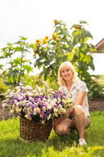 Positive Woman Sitting On The Grass On The Background Of A Bed With A Basket Of Dried Flowers In Her Hands And Posing For The Camera With A Smile On Her Face. Happy Woman Sitting With Grown Flowers