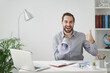 © ViDi Studio - Cheerful young business man in gray shirt sit at desk work on laptop pc computer in light office on white wall background. Achievement business career concept. Screaming in megaphone showing thumb up.