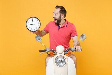 Wall Mural - Excited young bearded man guy in casual summer clothes driving moped isolated on yellow wall background studio portrait. Driving motorbike transportation concept. Mock up copy space. Holding clock.