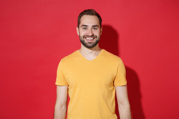 Smiling handsome young bearded man guy in casual yellow t-shirt posing isolated on red wall background studio portrait. People sincere emotions lifestyle concept. Mock up copy space. Looking camera.