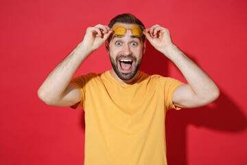 Wall Mural - Excited young bearded man guy in casual yellow t-shirt glasses posing isolated on red background studio portrait. People sincere emotions lifestyle concept. Mock up copy space. Keeping mouth open.