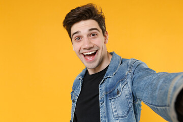 Close up of excited young man guy 20s in casual denim jacket posing isolated on yellow wall background studio portrait. People lifestyle concept. Mock up copy space. Doing selfie shot on mobile phone.