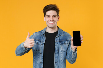 Smiling young man guy in casual denim jacket posing isolated on yellow background in studio. People lifestyle concept. Mock up copy space. Hold mobile phone with blank empty screen, showing thumb up.