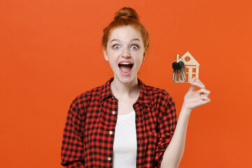 Shocked young readhead girl in casual red checkered shirt posing isolated on orange wall background studio portrait. People emotions lifestyle concept. Mock up copy space. Hold house bunch of keys.