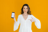 Fototapeta Na ścianę - Happy beautiful young woman with long curly hair holding blank screen mobile phone and pointing finger over yellow background