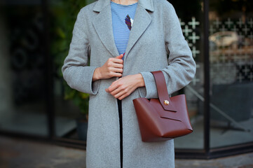 Fashionable young woman wearing grey coat, striped vest blouse, pants, holding brown bag . Street style.