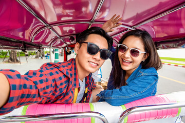 Cheerful young Asian couple tourists taking selfie while traveling on local colorful Tuk Tuk taxi exploring Bangkok city, Thailand