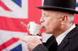 Englishman drinking afternoon tea from a cup and saucer, he's wearing a dark business suit with a bowler hat and standing against a Union Jack flag background.