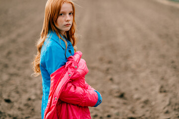 Beautiful strange red head girl in long blue coat and pink warm jacket spends a lot of time on the big empty field.