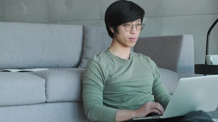 Canvas Print - A calm young asian man wearing eyeglasses is using his laptop sitting in the living room at home