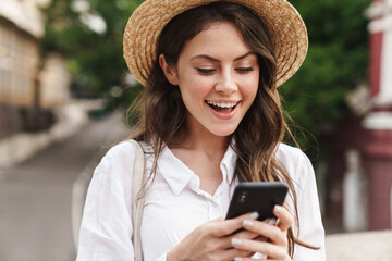 Wall Mural - Portrait of cheerful woman in straw hat using cellphone and smiling