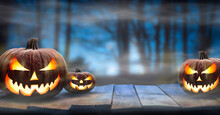 Three Spooky Halloween Pumpkins, Jack O Lantern, With Evil Face And Eyes On A Wooden Bench, Table With A Misty Night Forest Background With Space For Product Placement.